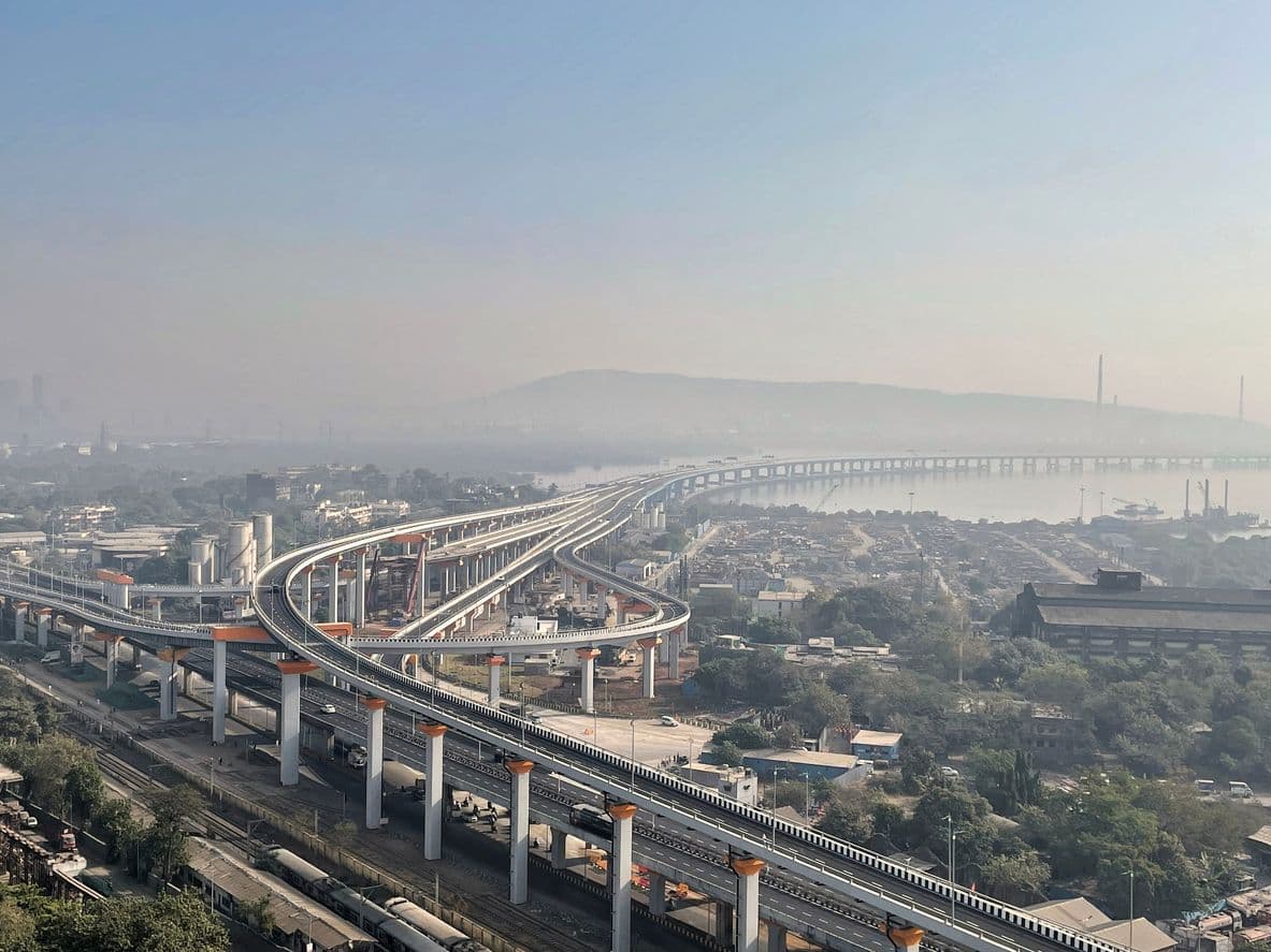 An aerial view of the multi-lane Sewri interchange of the Atal Setu bridge curving over the coastline and urban landscape in Mumbai during a hazy day.