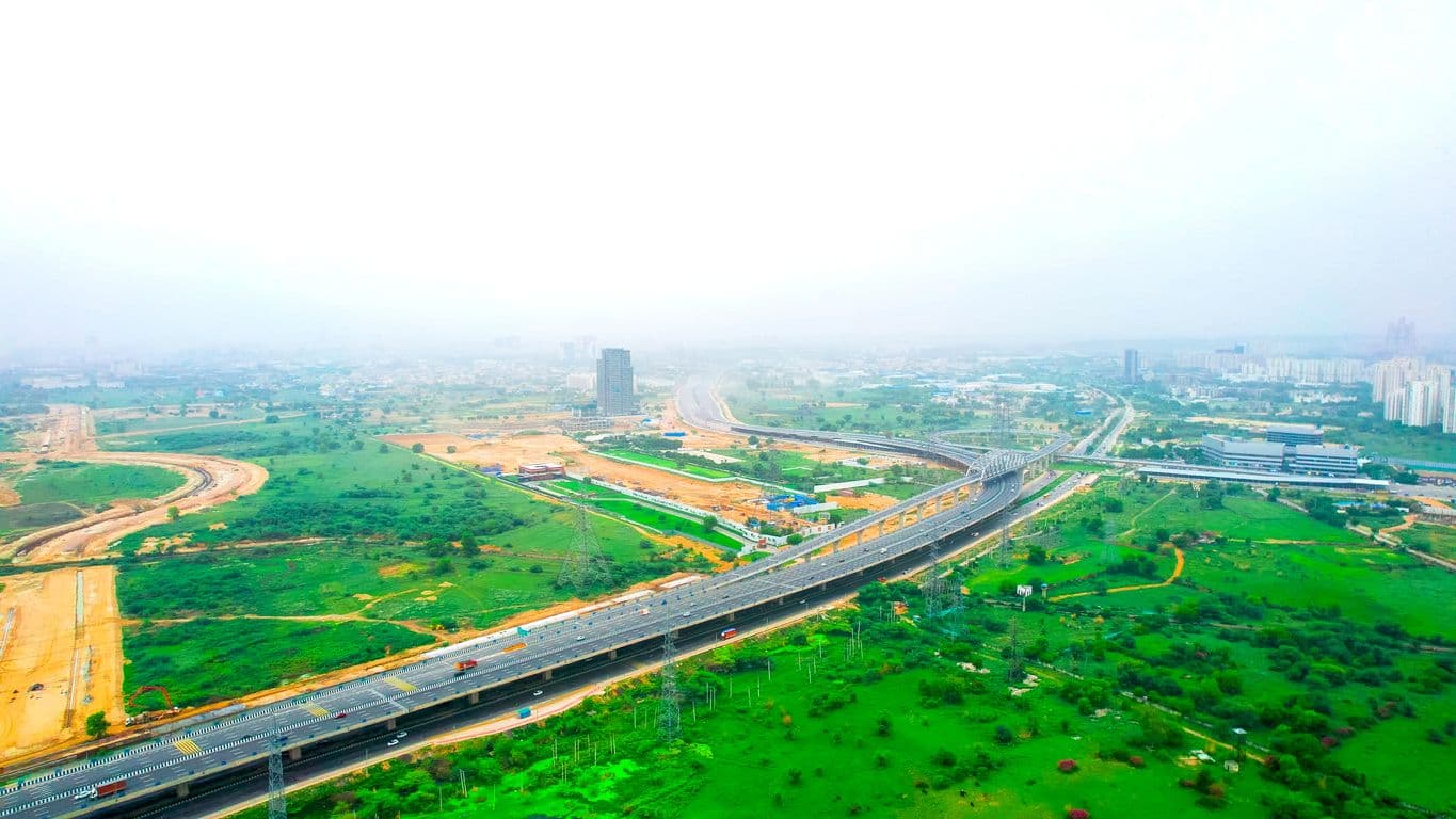 An aerial shot of a multi-lane elevated highway stretching across a green landscape. A massive arched steel bridge carries the expressway over a railway line, with extensive earthworks and under-construction ramps visible alongside it.