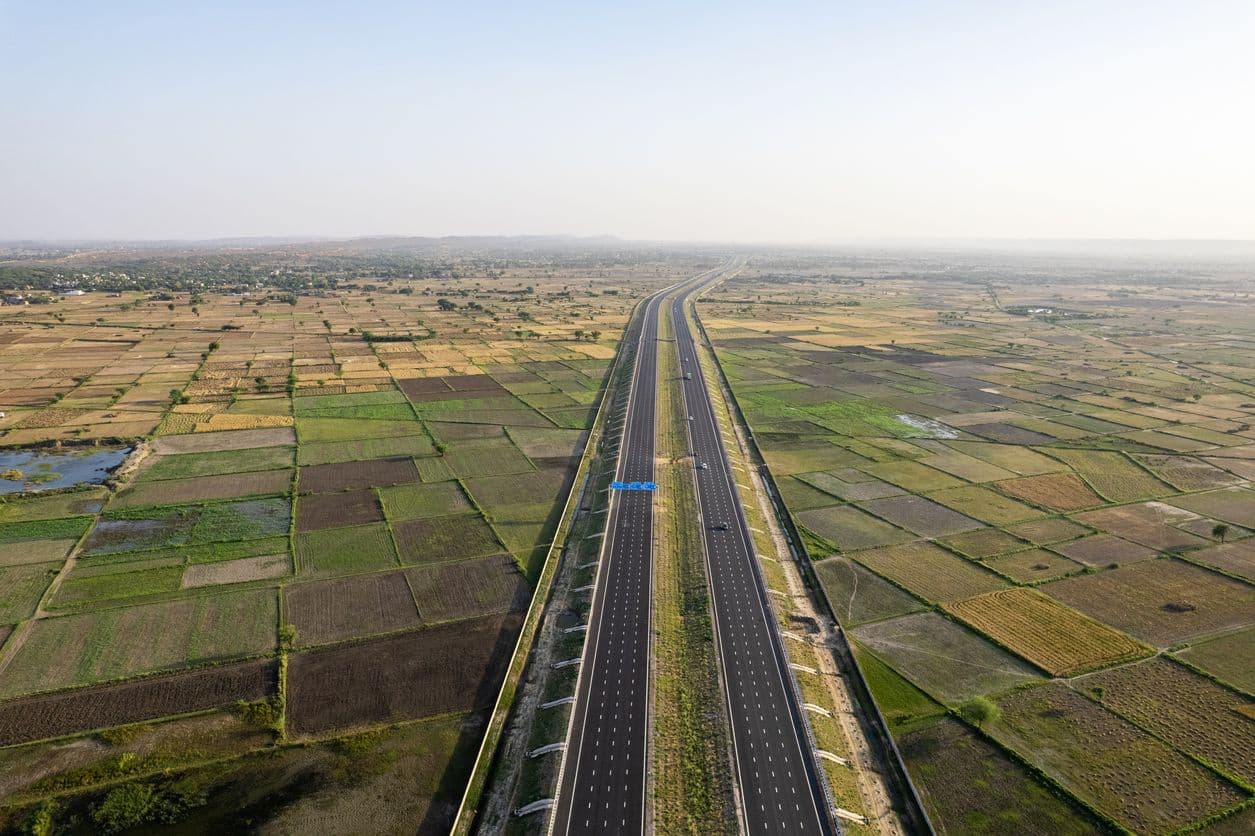 An aerial view of the wide, multi-lane Delhi-Mumbai Expressway cutting straight through a vast patchwork of green and brown fields.