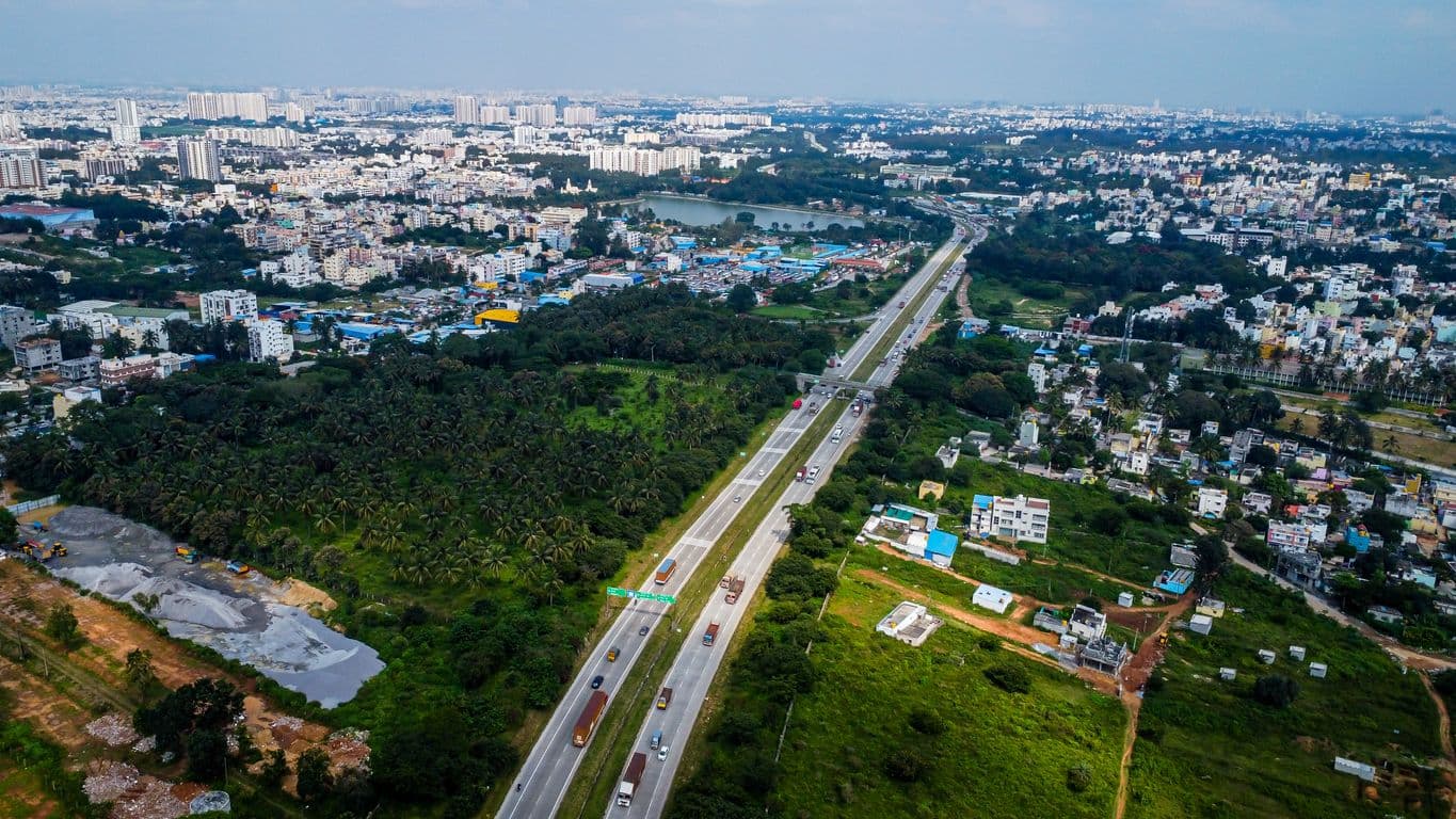 A high-altitude shot looking down the straight, wide Bengaluru-Mysuru Expressway. The highway cuts directly between dense urban housing on the left and open agricultural green spaces on the right.