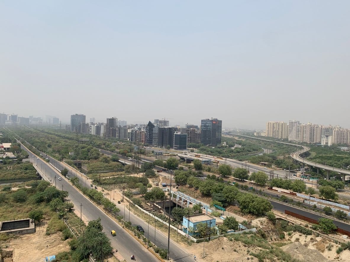An elevated view showing the concrete viaducts of the Noida Metro Aqua Line curving alongside the wide Noida-Greater Noida Expressway. Dense clusters of high-rise commercial and residential buildings form the skyline in the hazy background.