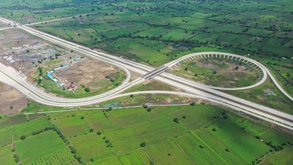 A top-down aerial view of a massive highway interchange constructed in the shape of a perfectly symmetrical double-loop or figure-eight. The grey concrete roads stand out starkly against the surrounding bright green agricultural fields