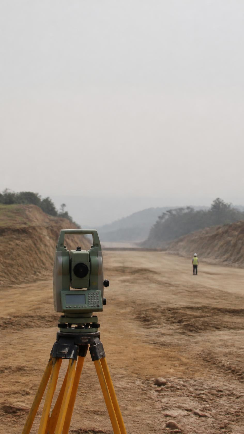 A survey total station on a tripod, an unfinished road corridor running away behind it.