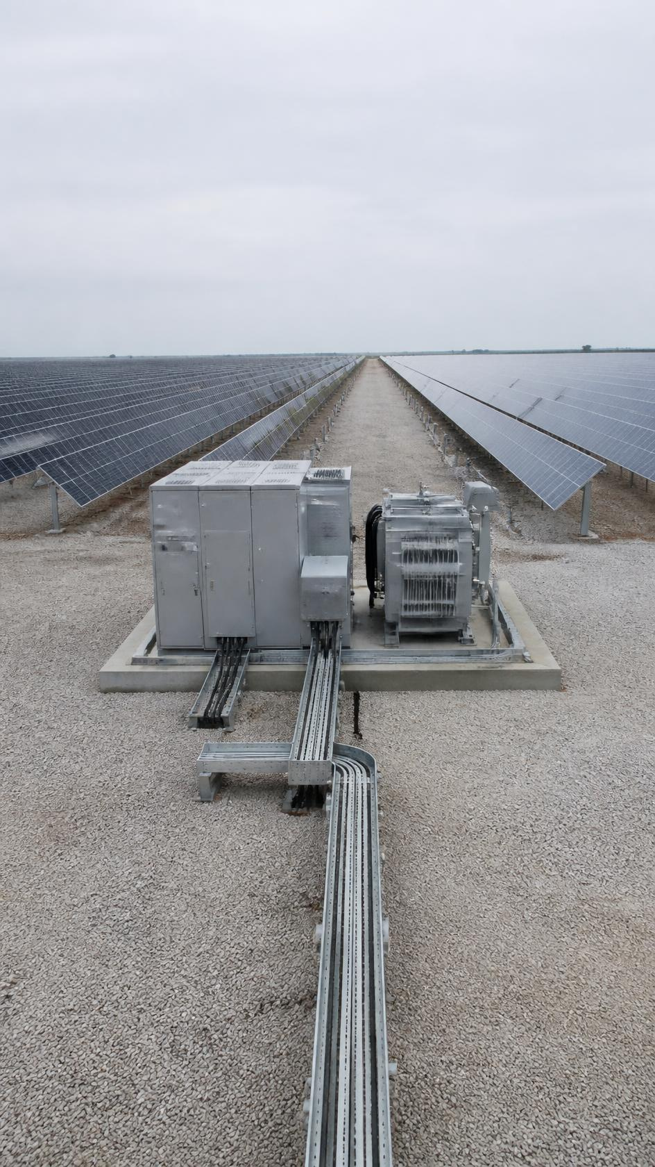 An inverter and transformer skid on gravel between rows of solar panels.